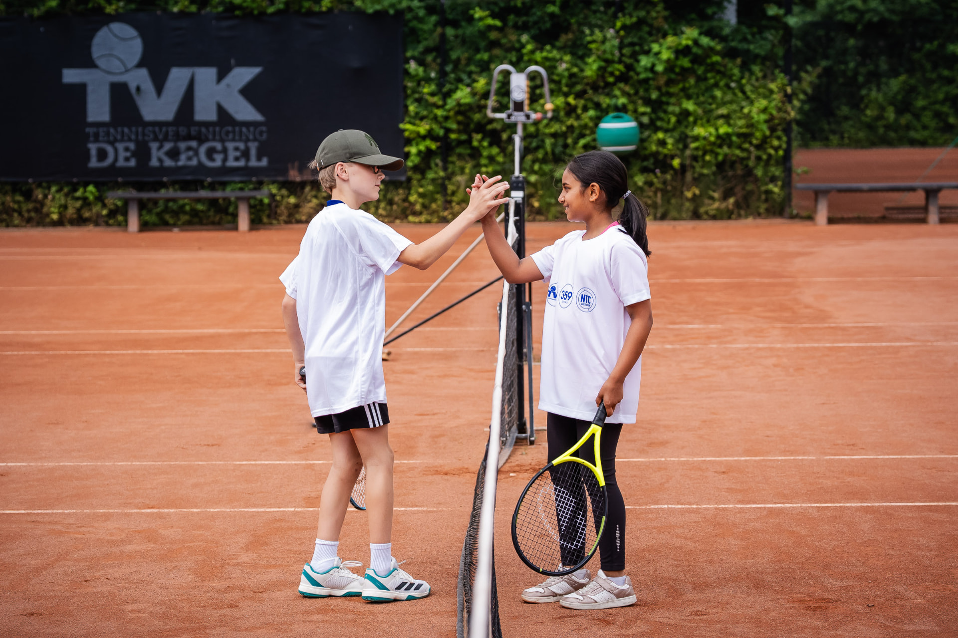 Twee kinderen geven elkaar een high five bij het net na een oefening op de gravelbaan — 359.tennis