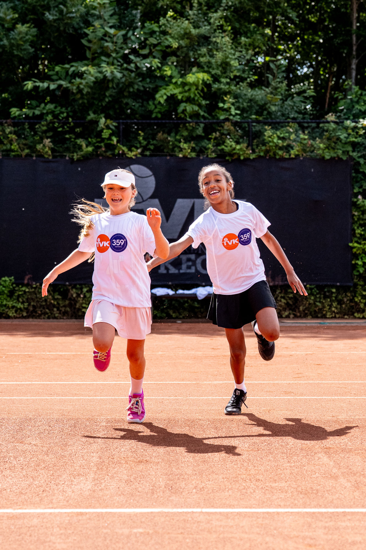 Twee meisjes rennen lachend samen over de gravelbaan tijdens tennistraining bij TV De Kegel — 359.tennis